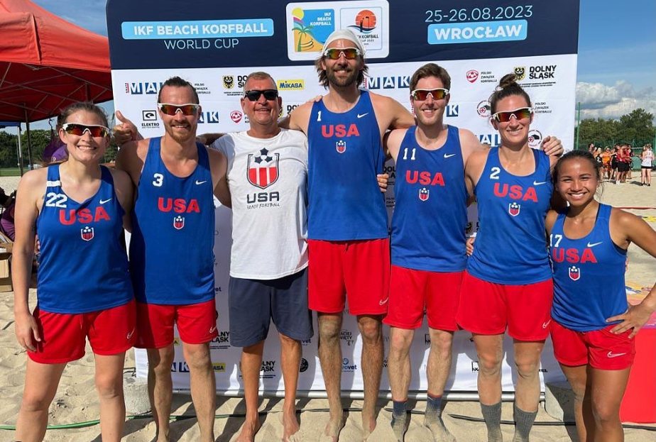The US Beach team wearing red and blue uniforms, standing in front of the IKF Beach Korfball Poland backdrop